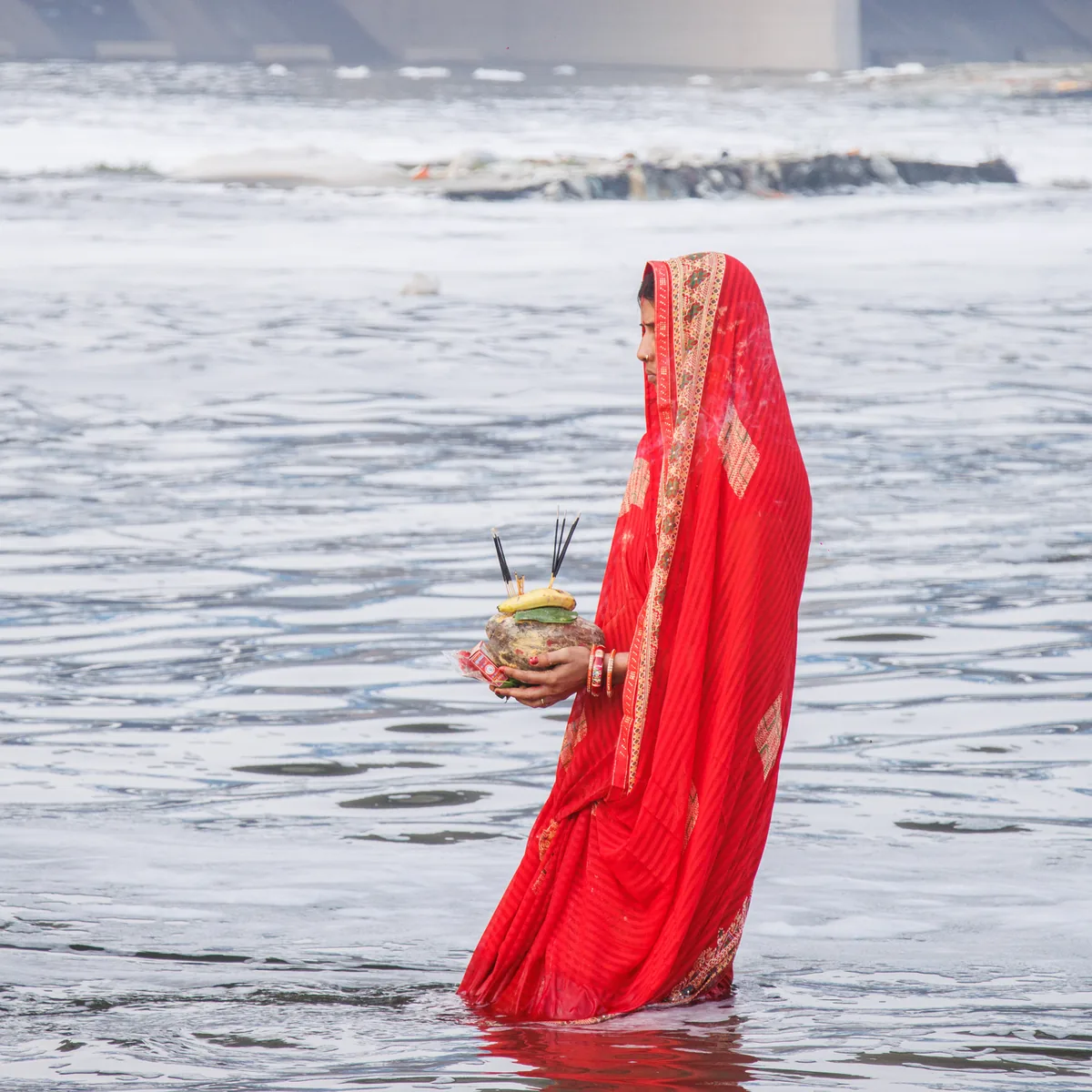 A woman in a red sari standing in a river, holding an offering to the Sun
